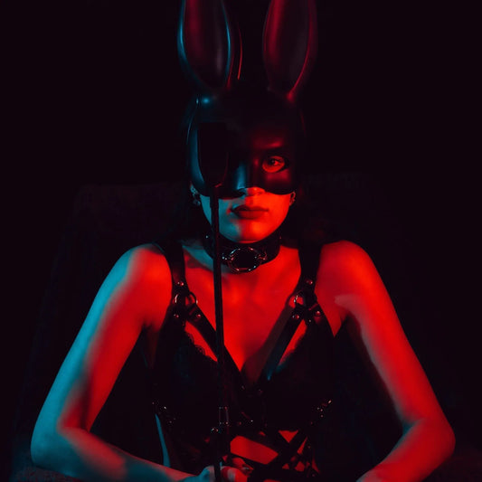 Woman in black bunny mask, leather harness, and leash, bathed in red and blue light