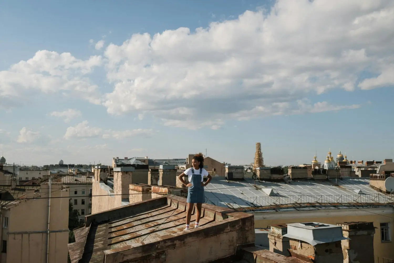 A young girl stands confidently on a city rooftop under a bright blue sky.